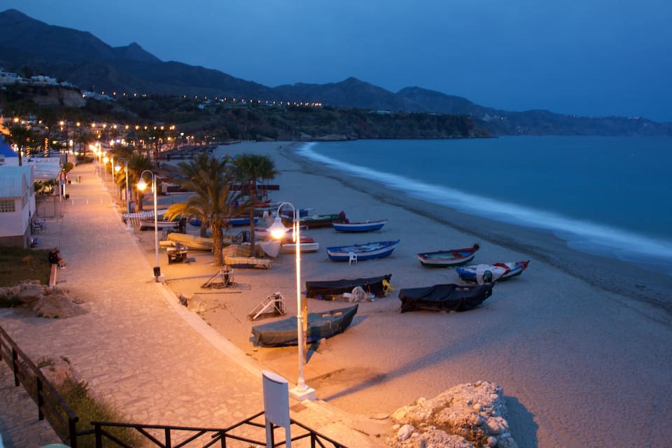 A beach at dusk with boats, a pathway, and a mountain in the background.