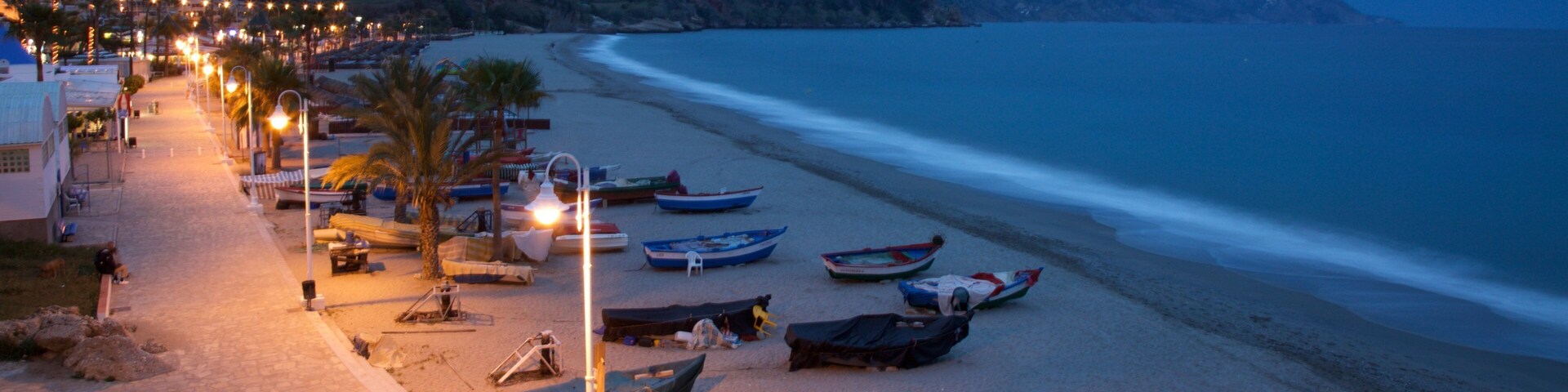 Una playa al atardecer con barcos, un camino y una montaña al fondo.