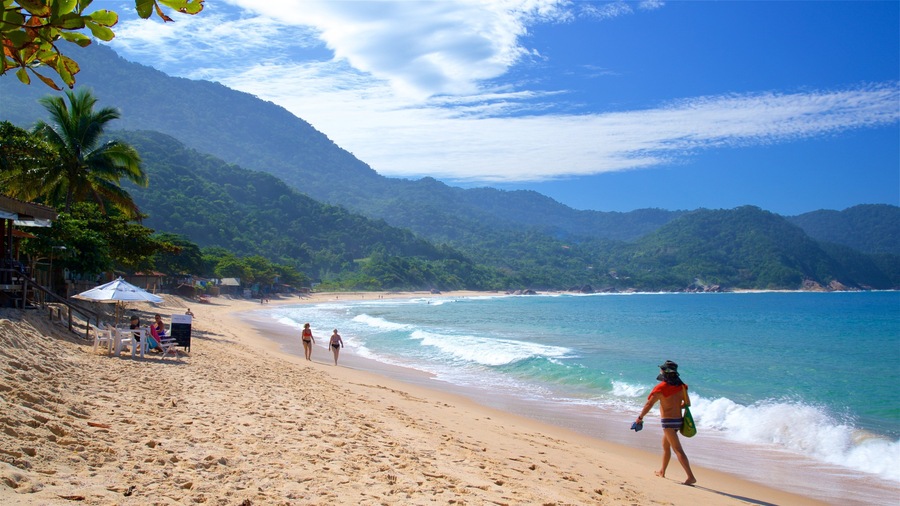 Uma praia com águas turquesa cristalinas, areal arenoso, palmeiras e montanhas ao fundo.