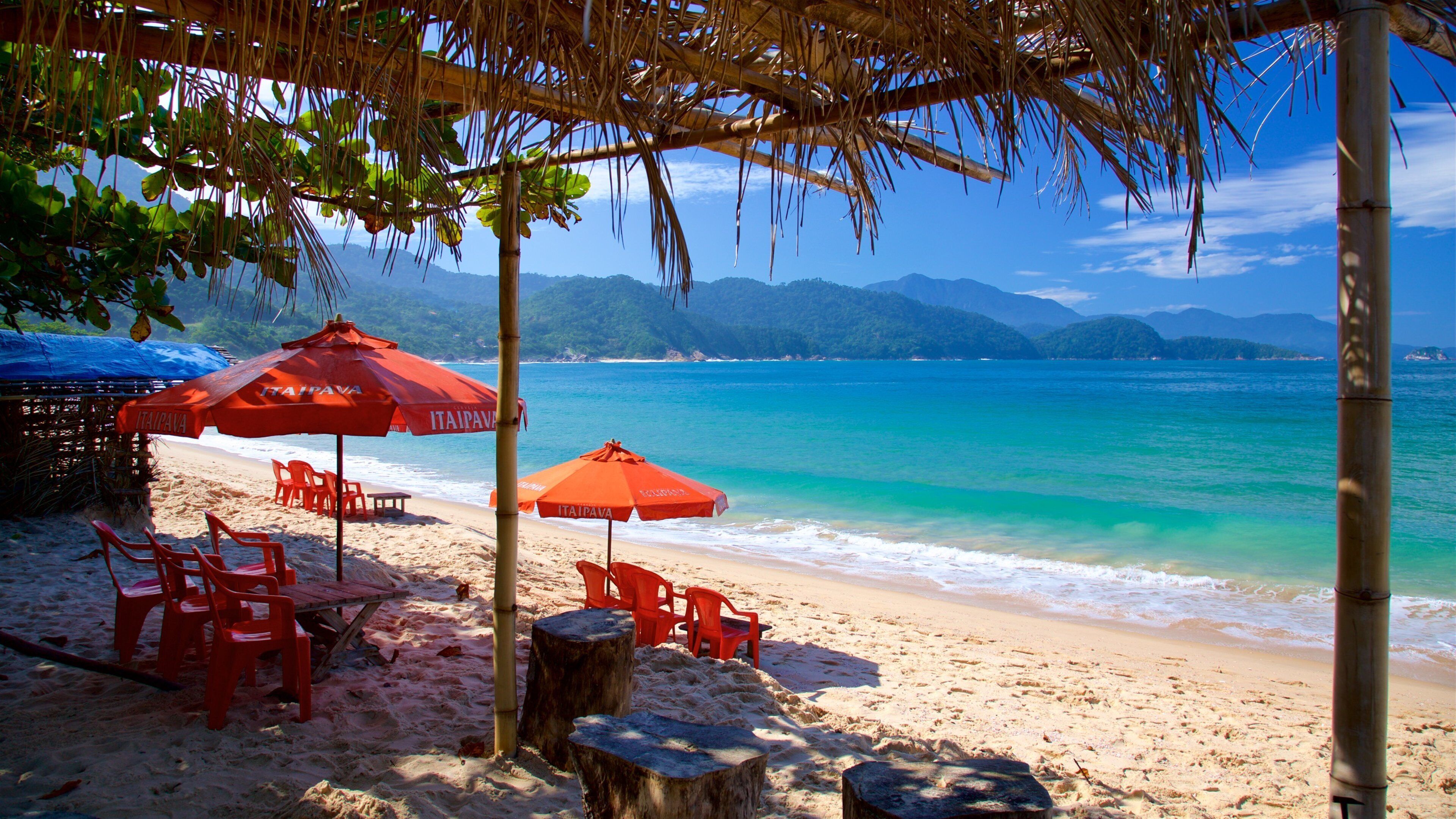 A beach with red umbrellas and chairs, a thatched roof, and a view of the ocean and mountains.