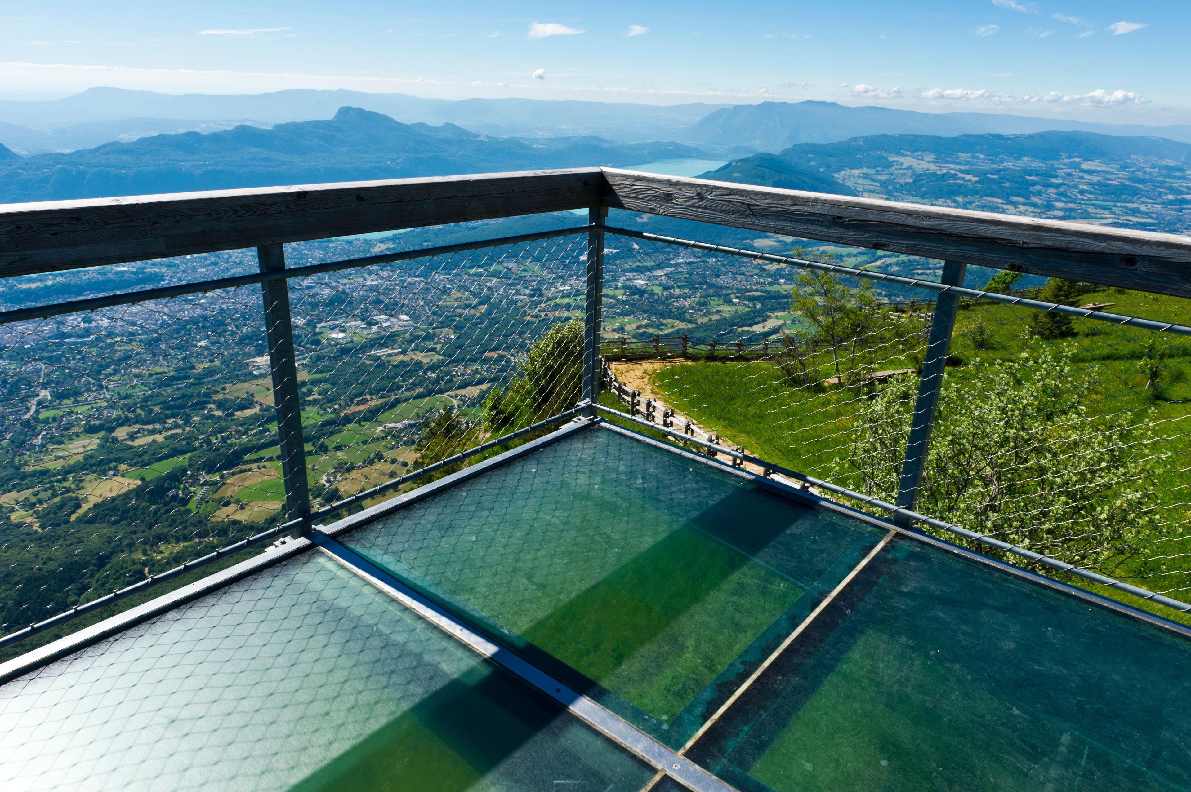 A glass-bottomed viewing platform with a metal railing overlooking a valley with mountains in the distance.