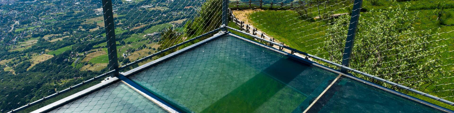A glass-bottomed viewing platform with a metal railing overlooking a valley with mountains in the distance.
