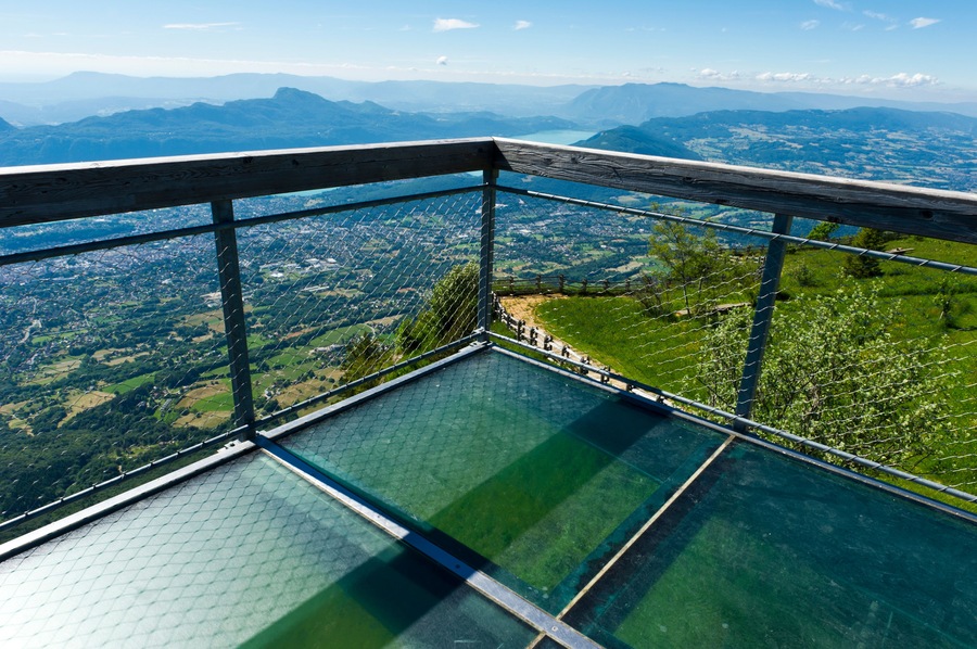 A glass-bottomed viewing platform with a metal railing overlooking a valley with mountains in the distance.
