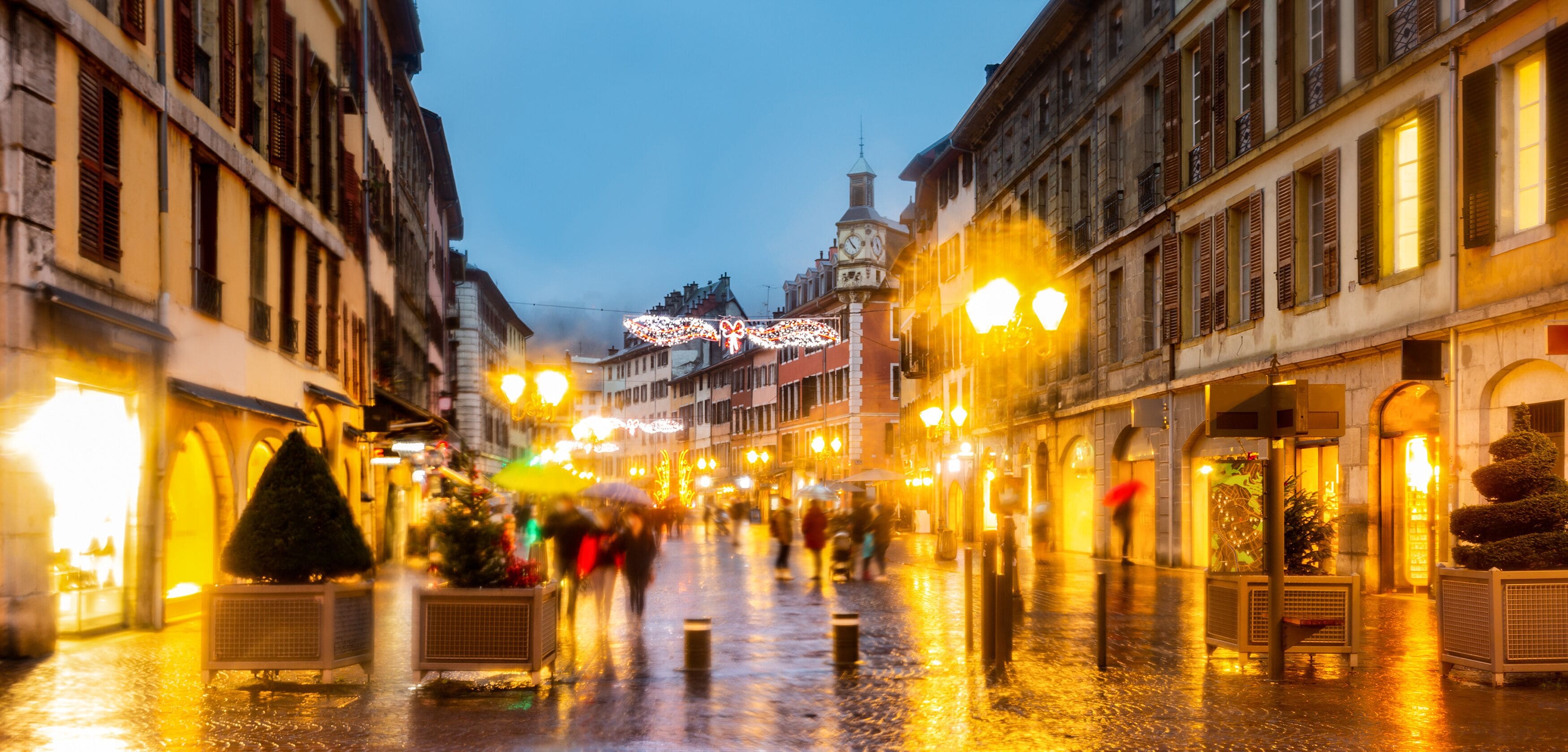 Uma rua da cidade molhada com edifícios históricos, uma torre do relógio e luzes festivas.