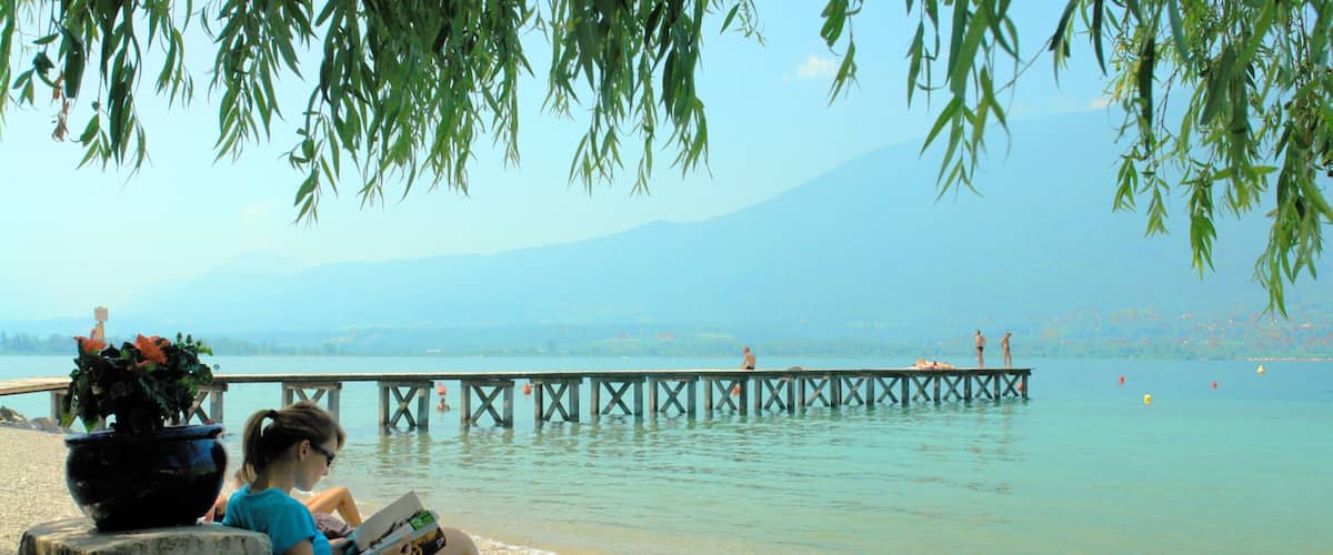 Eine Frau liest am See, eine Anlegestelle erstreckt sich ins Wasser und Berge sind im Hintergrund zu sehen.