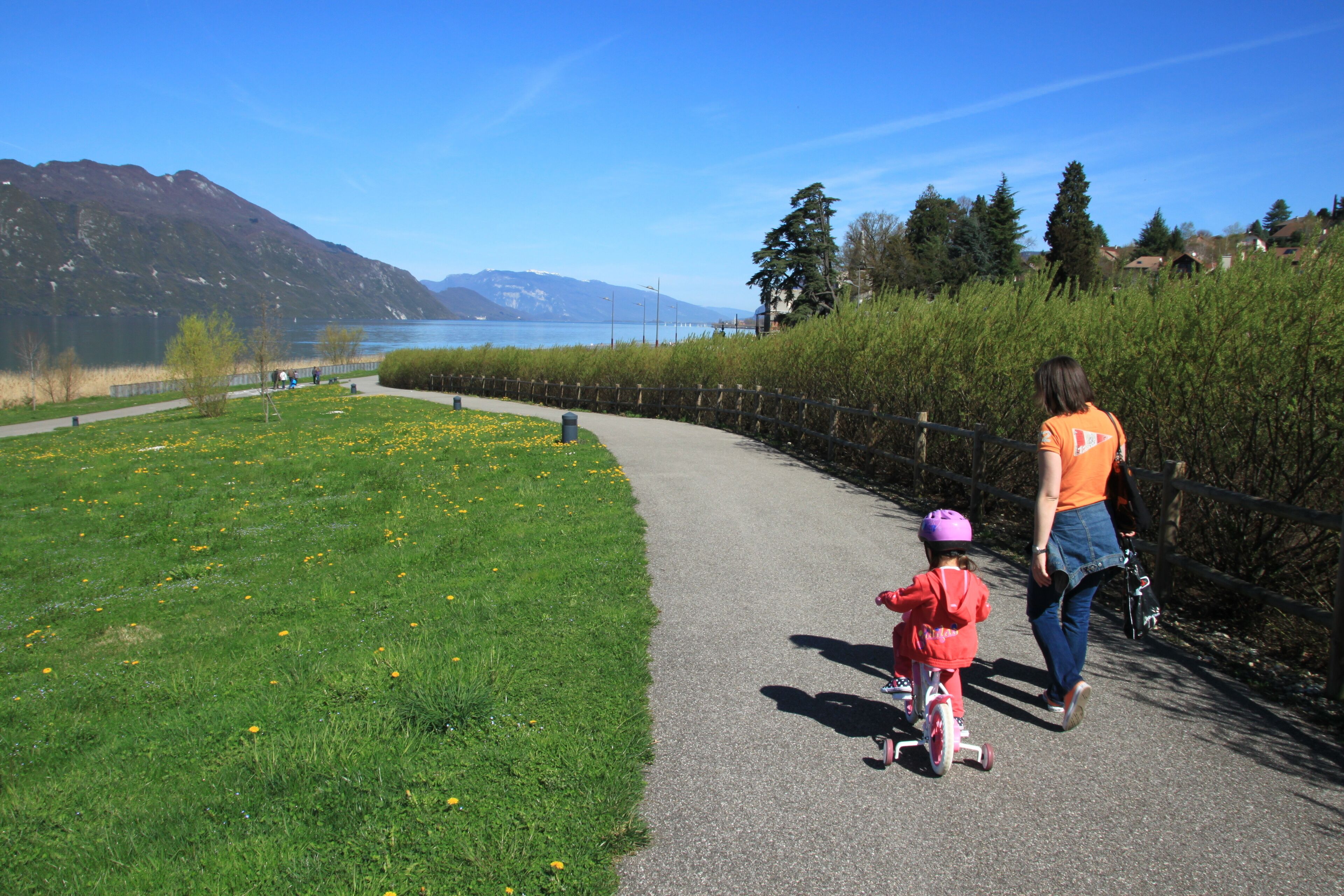 A paved path alongside a grassy area with a lake and mountains in the background.