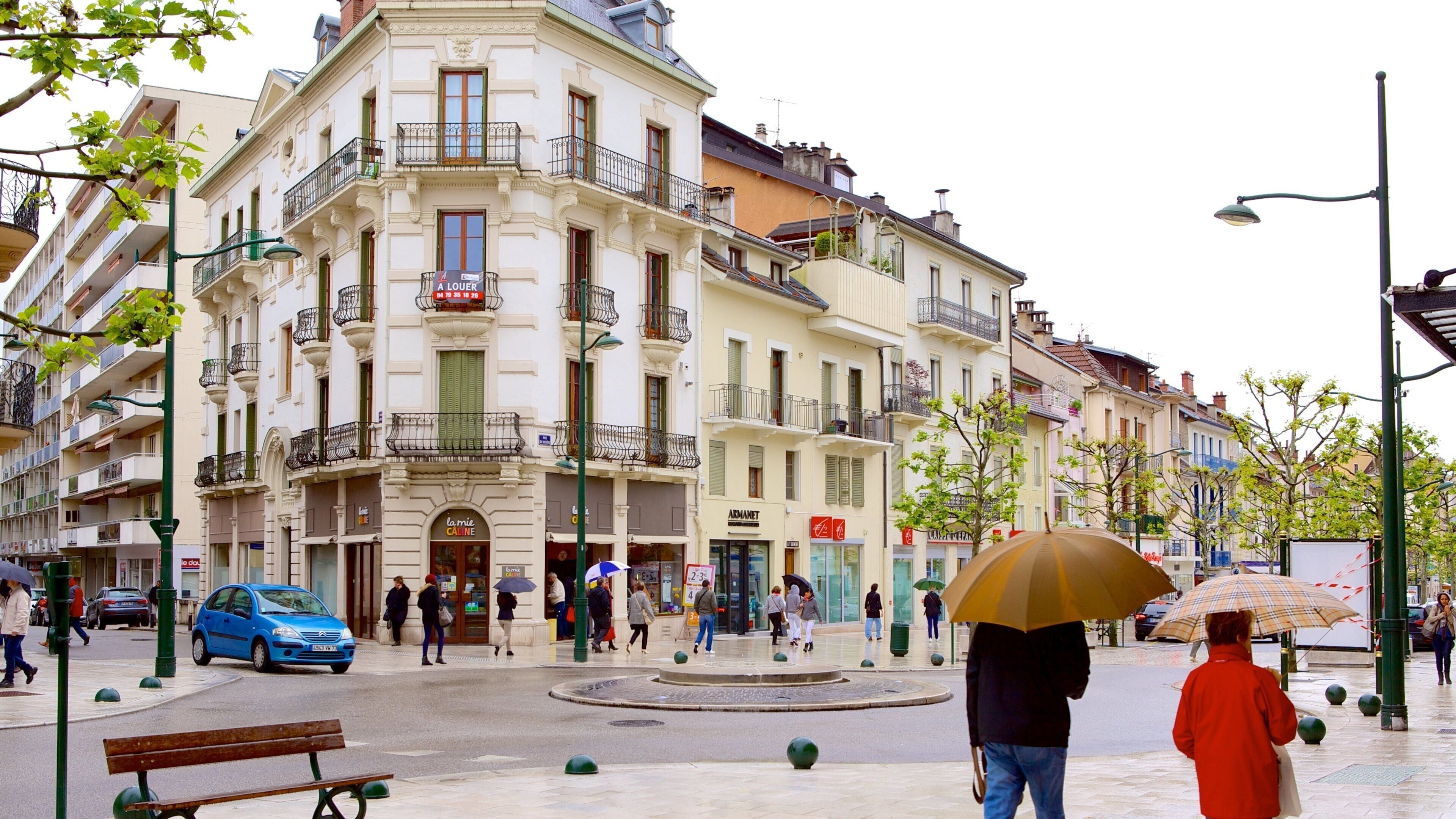 A European city square with historic buildings, a blue car, and people walking.