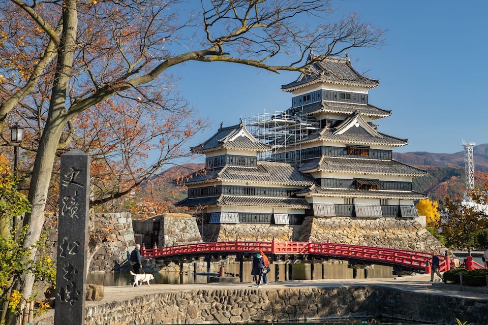Un castillo japonés tradicional con varios niveles, rodeado de follaje otoñal y un puente de piedra.