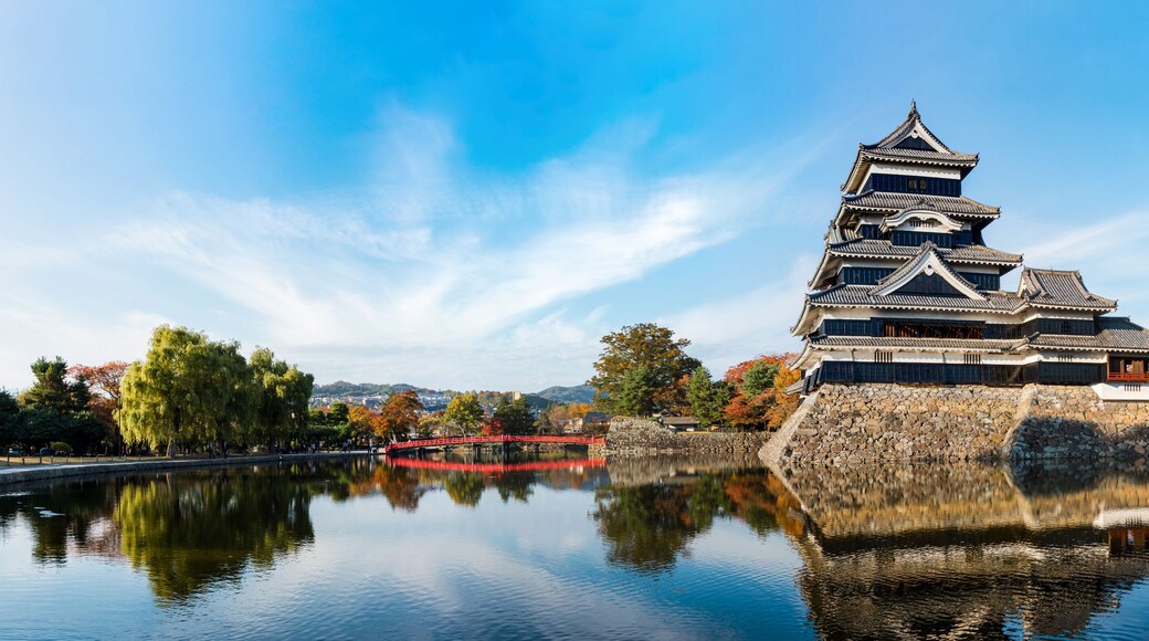 Eine traditionelle japanische Burg mit Spiegelbild im Wasser, umgeben von Herbstbäumen und blauem Himmel.