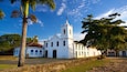 A white church with a bell tower, surrounded by palm trees and a clear blue sky.