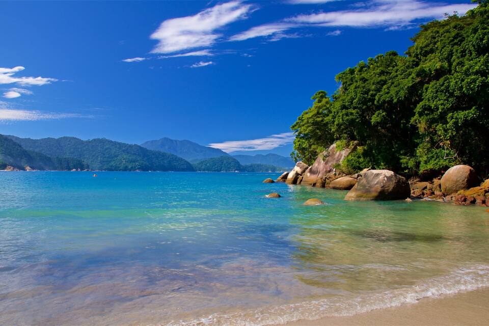 A clear turquoise sea, rocky shoreline, and lush green mountains under a blue sky.