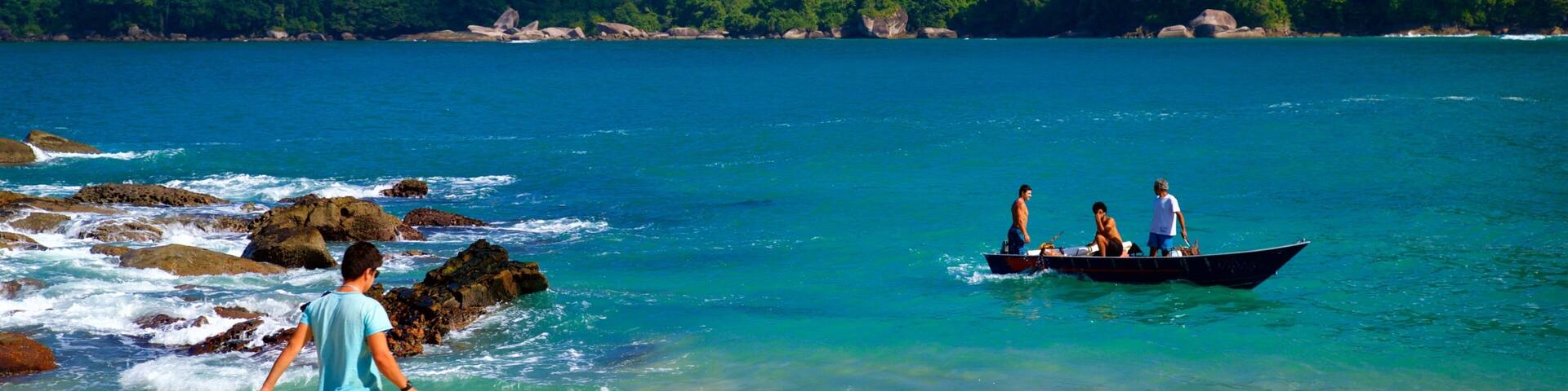 A beach with clear turquoise water, a rocky shoreline, and a boat with people in the distance.