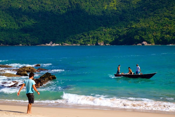 A beach with clear turquoise water, a rocky shoreline, and a boat with people in the distance.