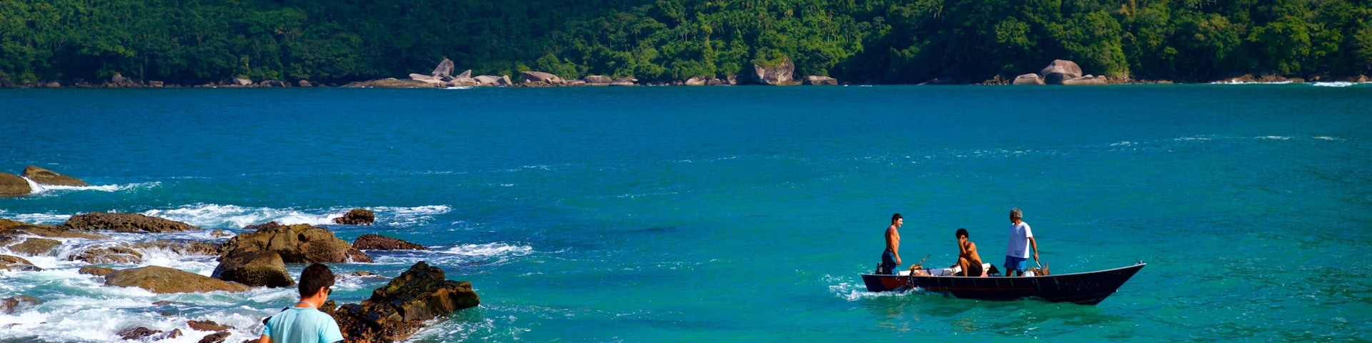 Una playa con agua turquesa cristalina, una costa rocosa y un barco con gente a lo lejos.