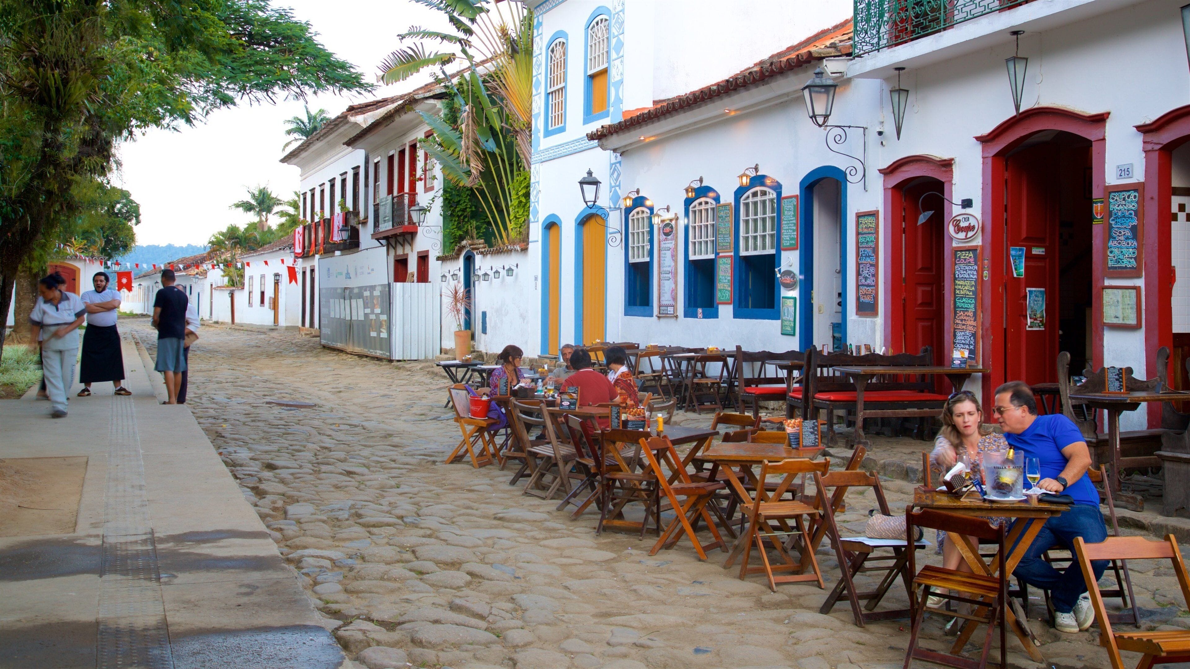 A cobblestone street lined with colorful buildings, outdoor seating at cafes, and people walking.