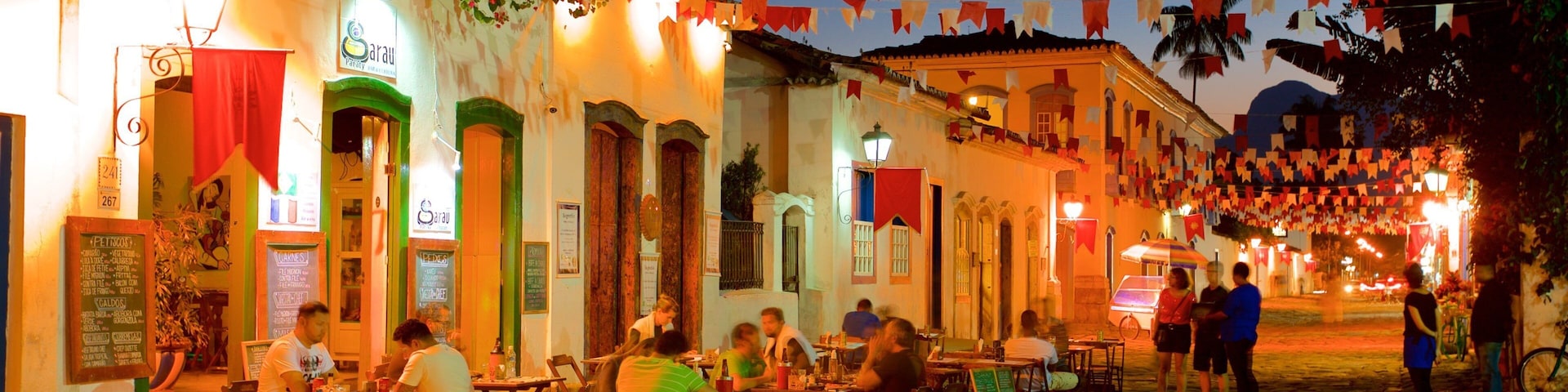 Ruas de paralelepípedos à noite, com mesas de café ao ar livre, decoradas com bandeiras coloridas.
