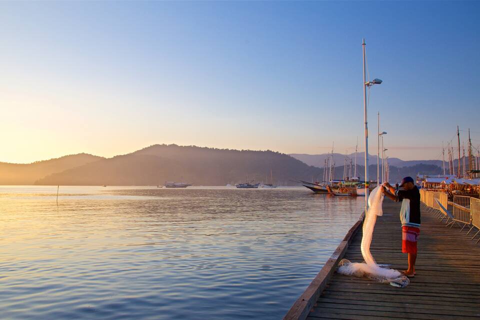 A wooden pier extending into calm waters with boats and a mountainous backdrop during sunset.