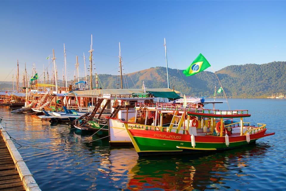 A dock with colorful boats, a Brazilian flag, and a mountainous landscape in the background.