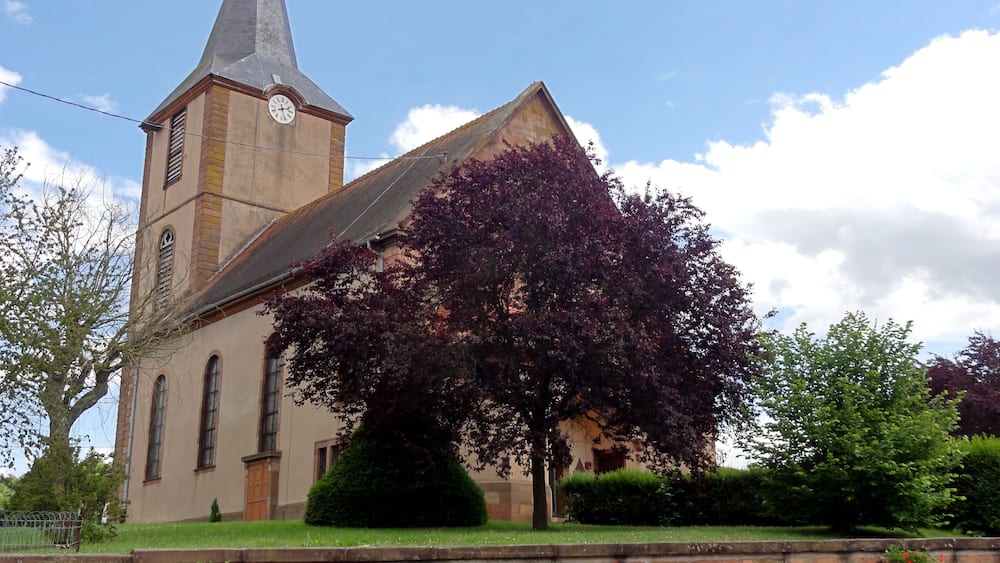 Une église dotée d’un clocher imposant et d’une horloge, entourée d’arbres et d’un mur en briques.