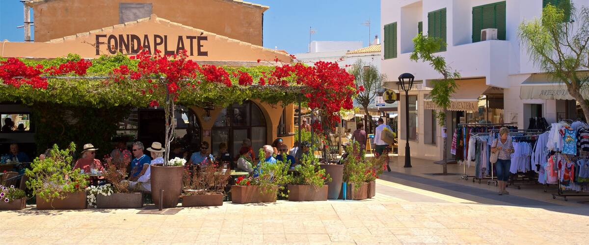 A sunny outdoor café with red flowers, people sitting and walking, and a clothing store in the background.