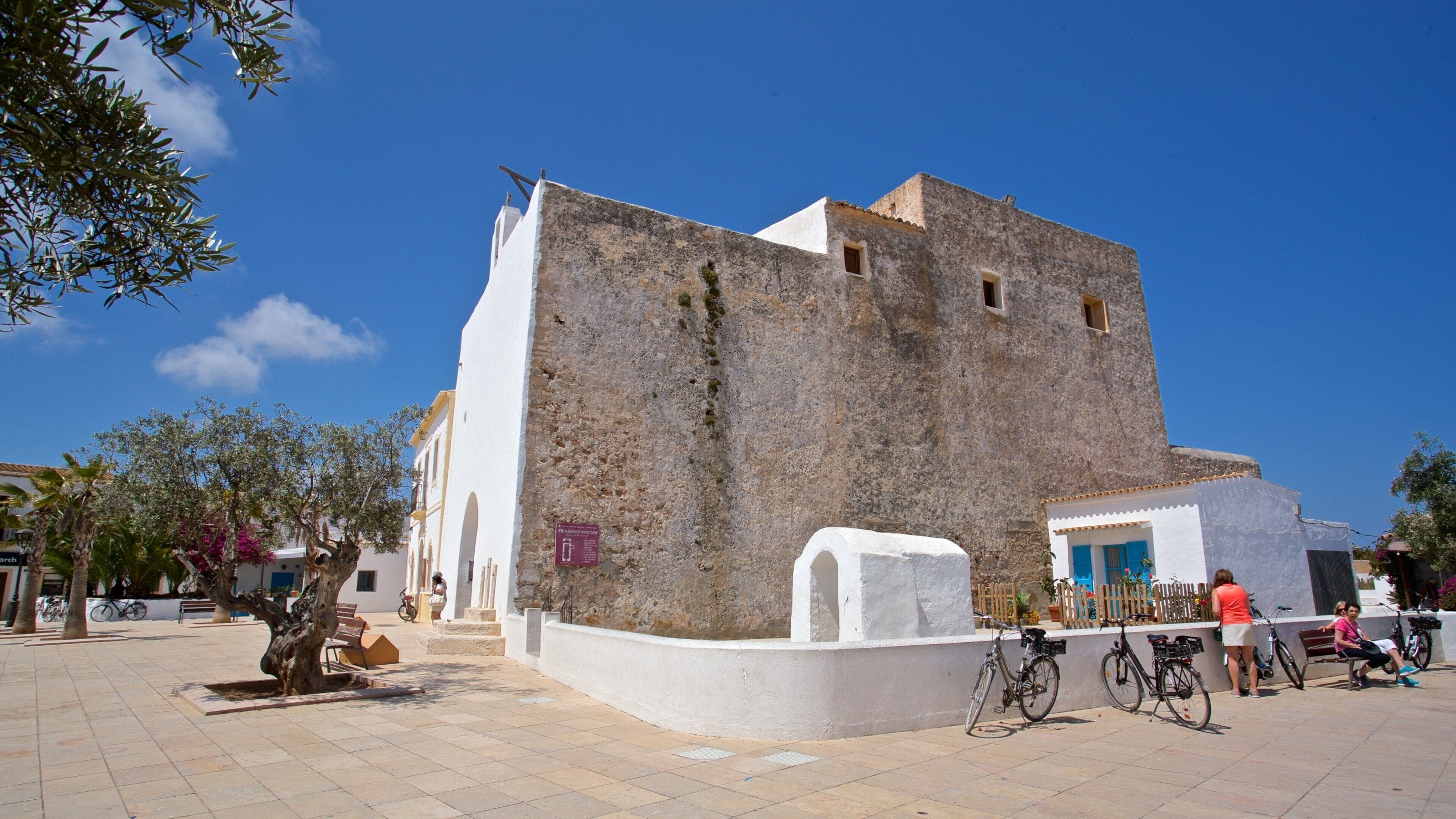 A historic building with a white facade and stone walls, surrounded by a paved square with benches and bicycles.
