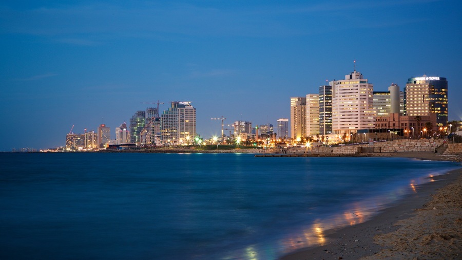A coastal cityscape at dusk with illuminated buildings and a calm sea.