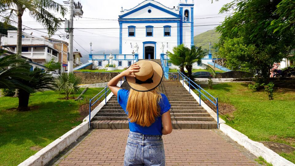 A woman in denim shorts and a blue shirt walking up a brick staircase towards a blue and white church with a bell tower.