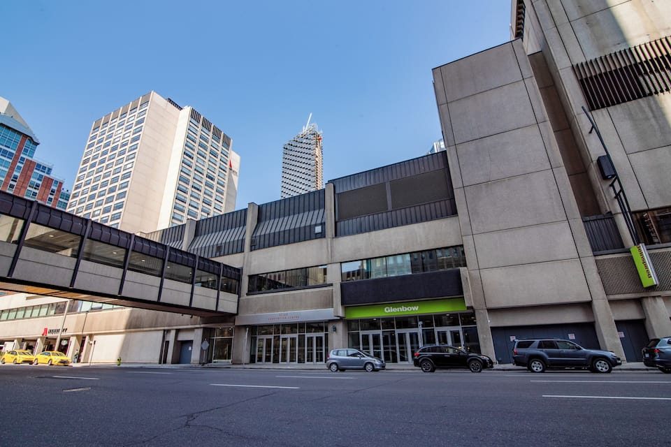 A modern urban street with a pedestrian bridge, a building with a 'Glenbow' sign, and several parked cars.