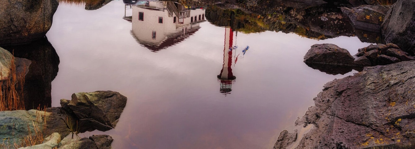 Um farol numa formação rochosa com uma casa por perto, refletido em águas calmas.