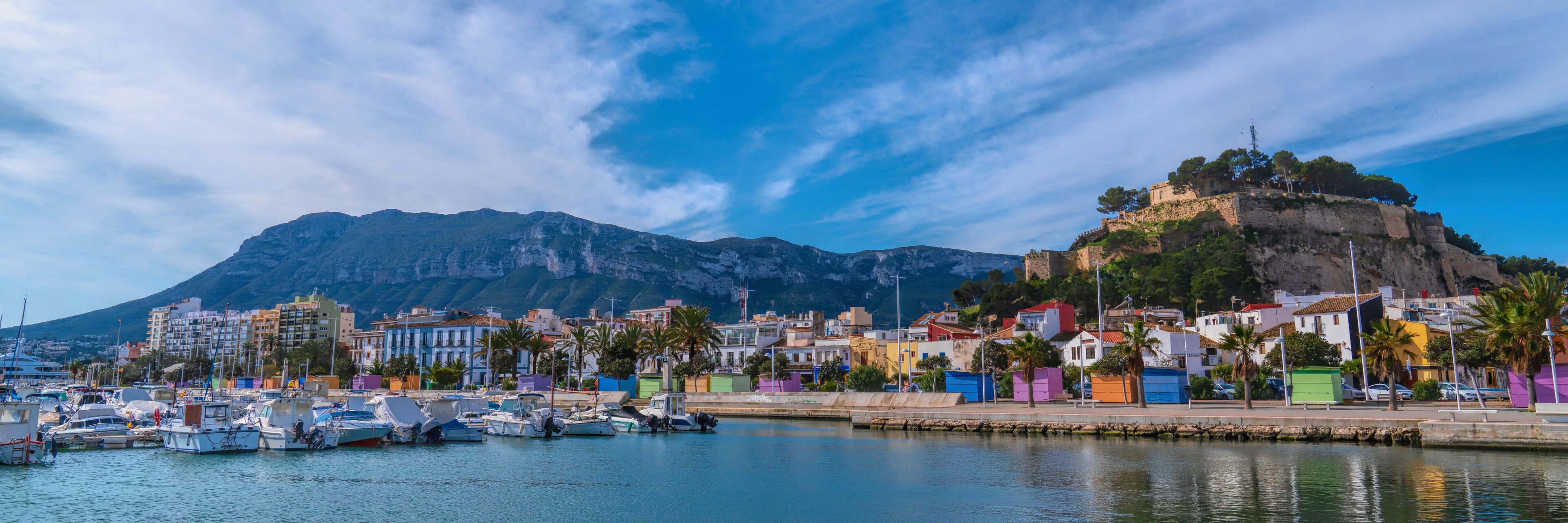 A coastal town with colorful buildings, a marina, and a prominent hill with ruins.