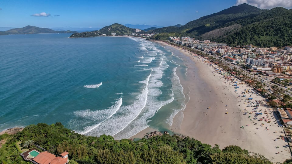 A coastal city with a long sandy beach, dense greenery, and mountains in the background.