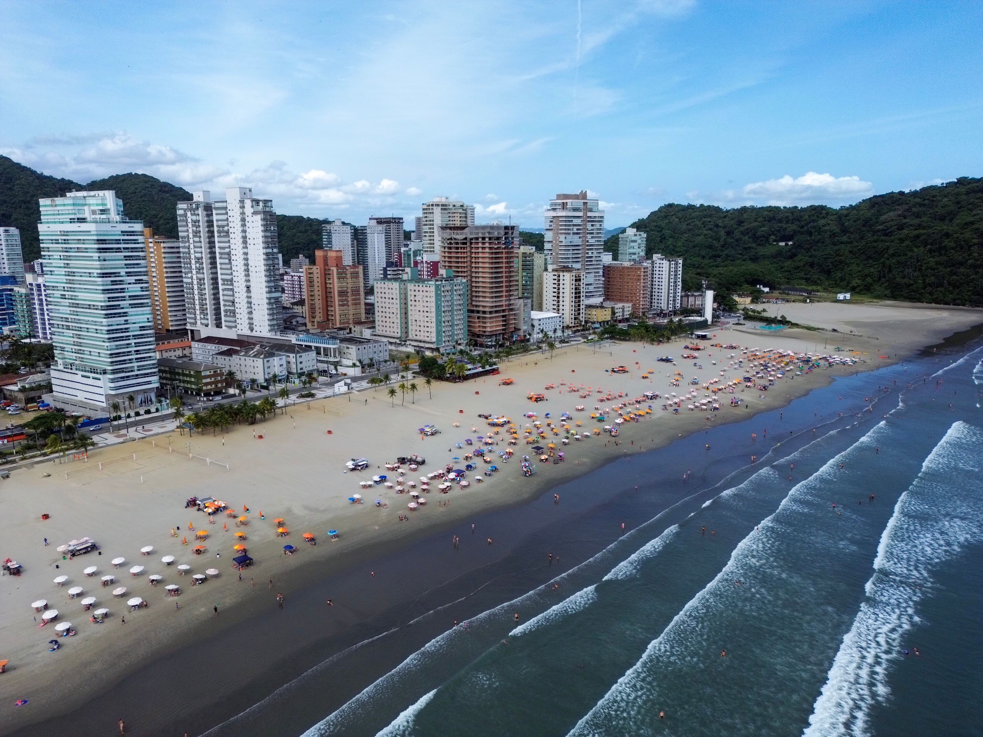 Aerial view of Praia Grande, Sao Paulo, Brazil, showing the coastline with waves reaching the sandy beach, high-rise buildings, and lush green hills in the background under a blue sky