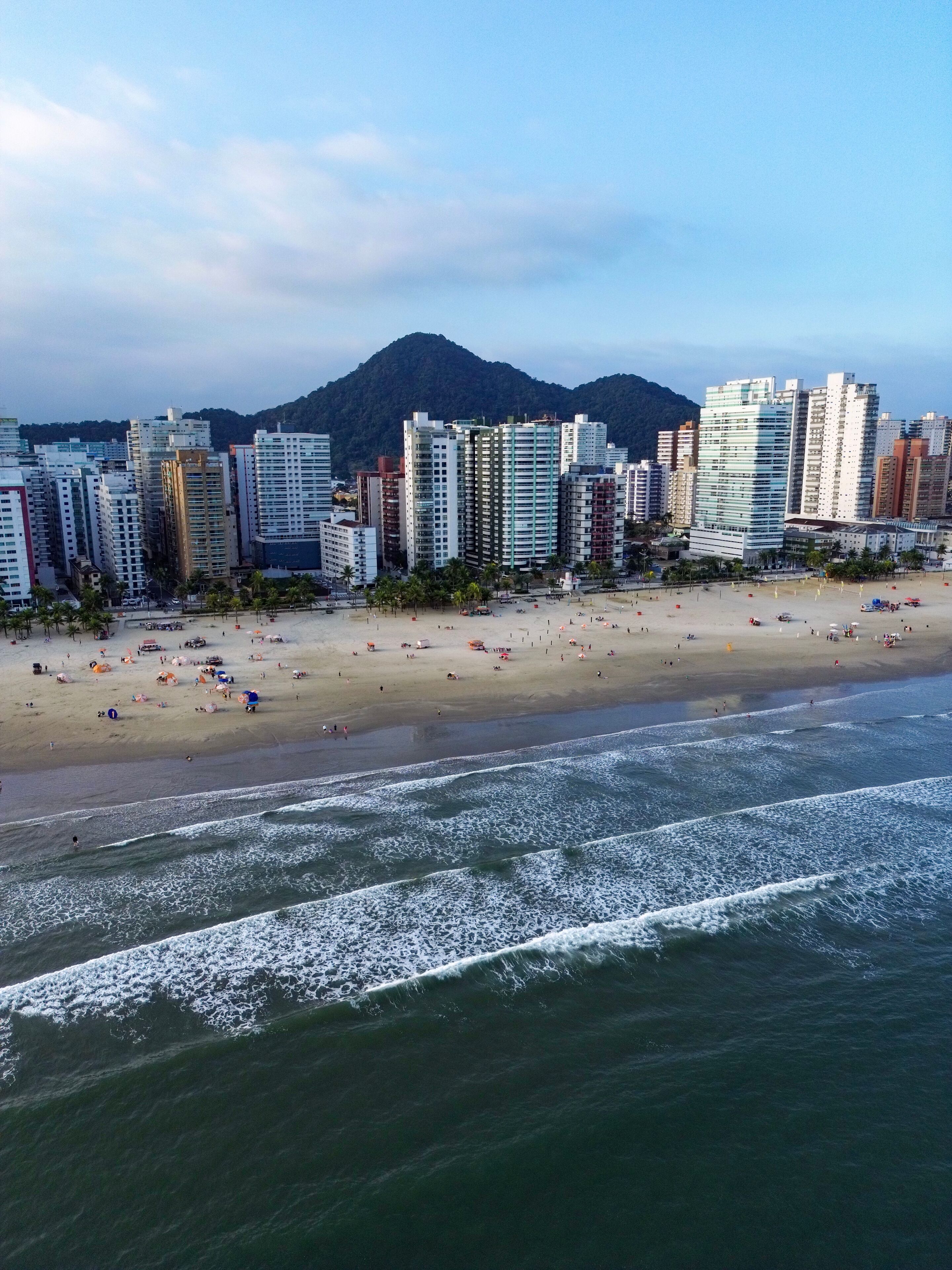 Aerial view of Praia Grande, Sao Paulo, Brazil, showing the coastline with waves reaching the sandy beach, high-rise buildings, and lush green hills in the background under a blue sky