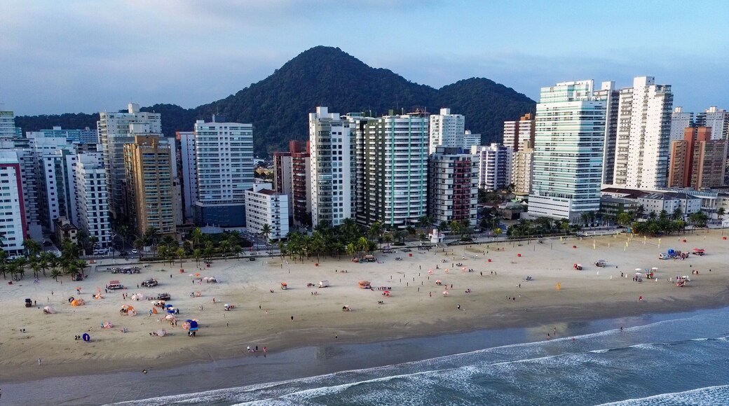 Aerial view of Praia Grande, Sao Paulo, Brazil, showing the coastline with waves reaching the sandy beach, high-rise buildings, and lush green hills in the background under a blue sky