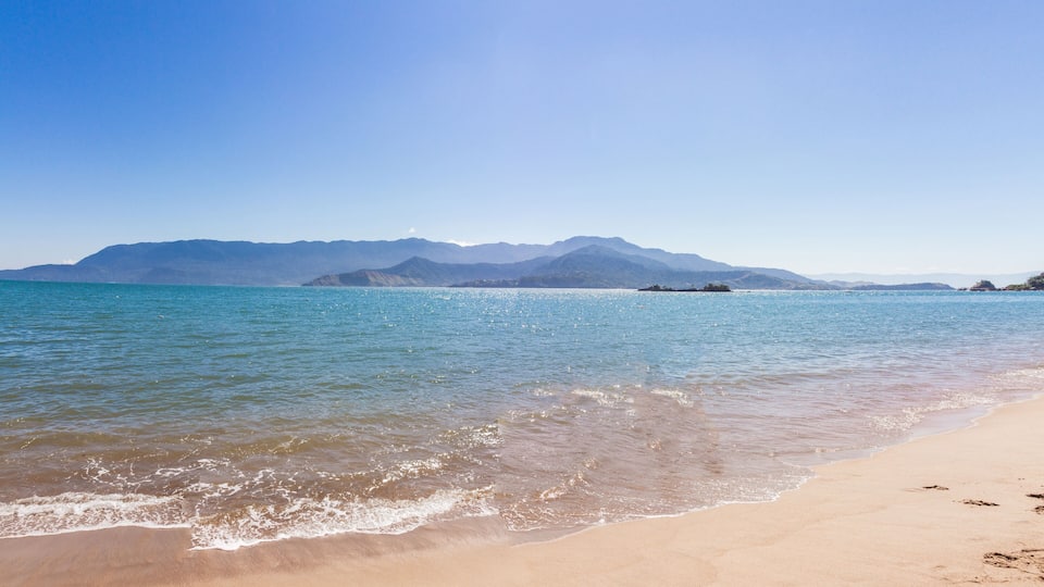 A beach with clear turquoise water, gentle waves, and distant mountains.
