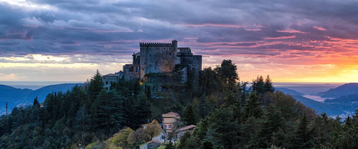A medieval castle on a hill surrounded by dense forest, with a sunset in the background.