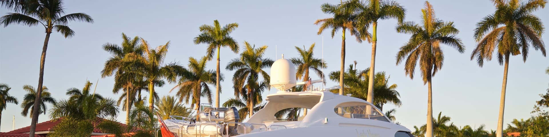 A large white yacht docked at a marina surrounded by palm trees and a building with a red roof.