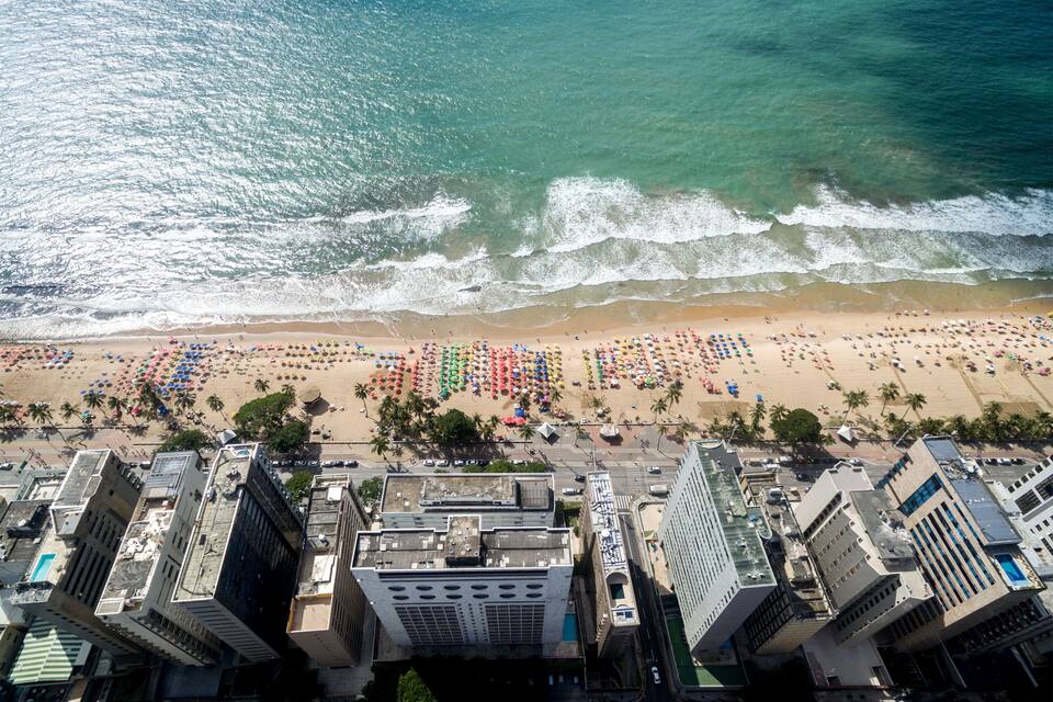 Vista aérea de una playa con sombrillas coloridas y palmeras, rodeada de edificios altos.