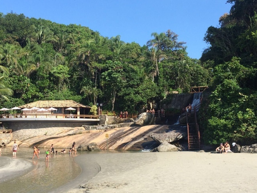 Een strand met een waterval, mensen die zwemmen en een constructie met rieten dak.