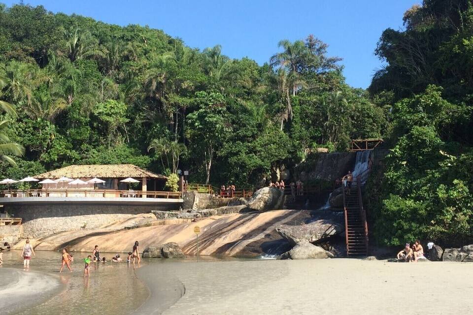 A beach with a waterfall, people swimming, and a thatched-roof structure.