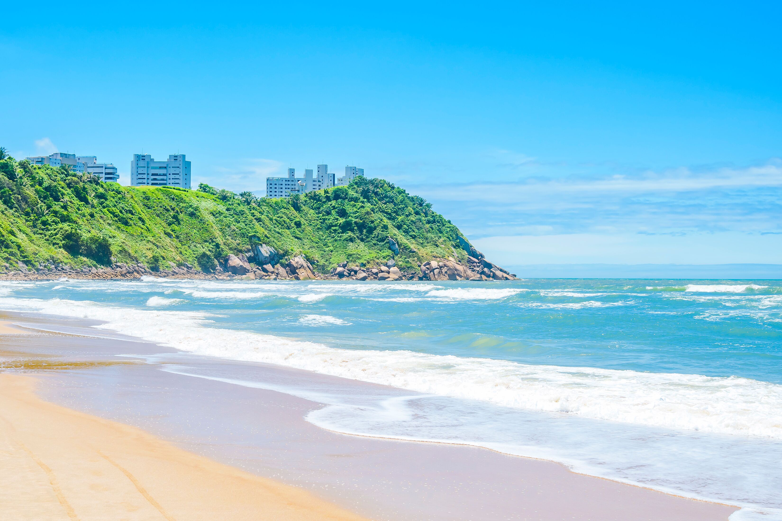 Beautiful Brazilian beach of the Paulista coast on a sunny day. The sea, some waves, the sand and a hill with some buildings on top. Photo at Praia do Tombo beach - Guaruja SP Brazil.