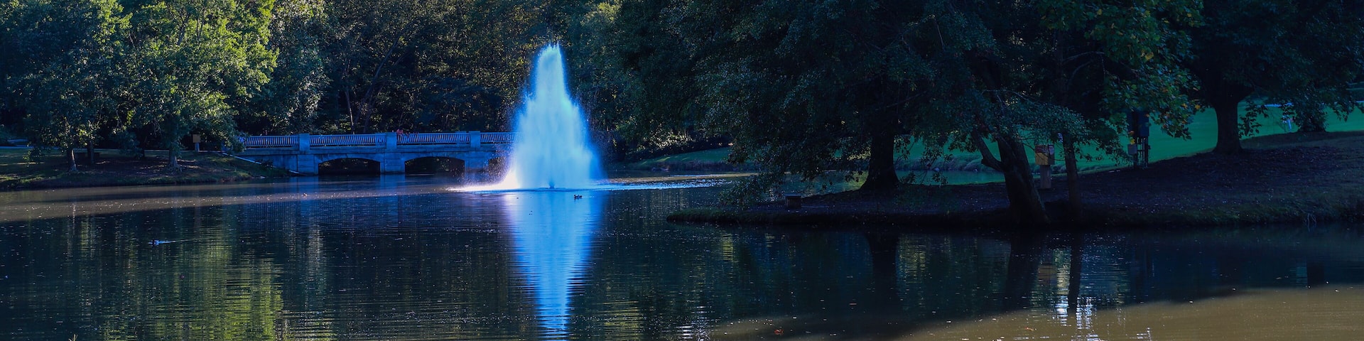 a gorgeous shot of the silky green lake water with a fountain in the middle of the lake with a stone bridge surrounded by lush green and autumn colored trees at Lenox Park in Brookhaven Georgia USA