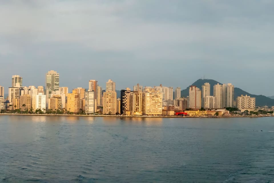 A city skyline with high-rise buildings along a waterfront, a cruise ship, and mountains in the background.