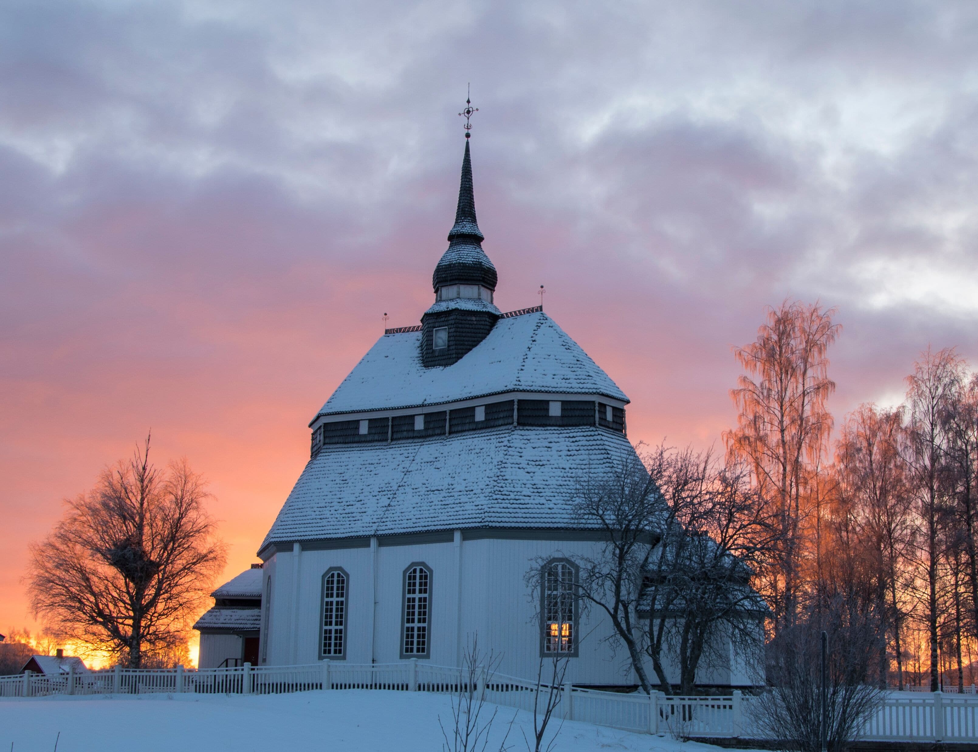 Una iglesia cubierta de nieve con una aguja, rodeada de árboles desprovistos de hojas y una cerca, durante la puesta de sol.