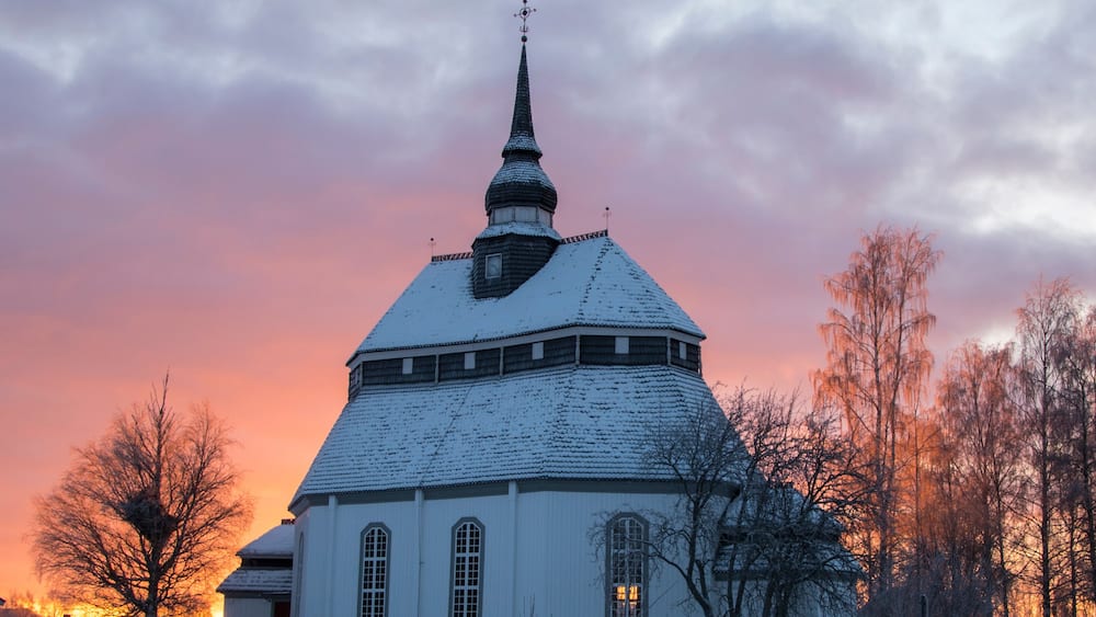 A snow-covered church with a spire, surrounded by bare trees and a fence, during sunset.