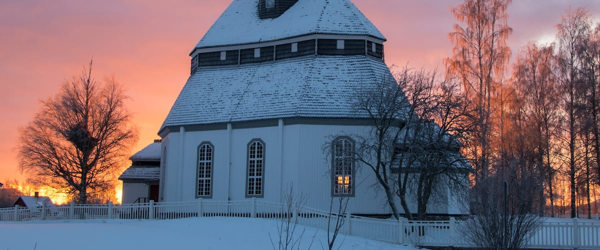 Une église enneigée, surmontée d’une flèche, entourée d’arbres sans feuilles et d’une clôture, au coucher du soleil.