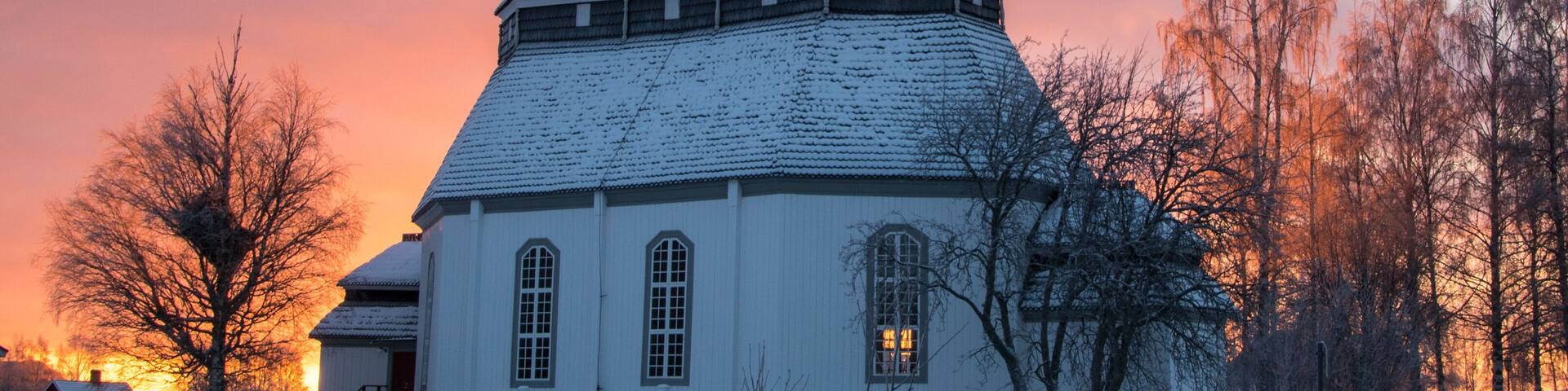 A snow-covered church with a spire, surrounded by bare trees and a fence, during sunset.