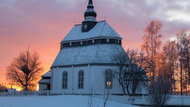 Una iglesia cubierta de nieve con una aguja, rodeada de árboles desprovistos de hojas y una cerca, durante la puesta de sol.
