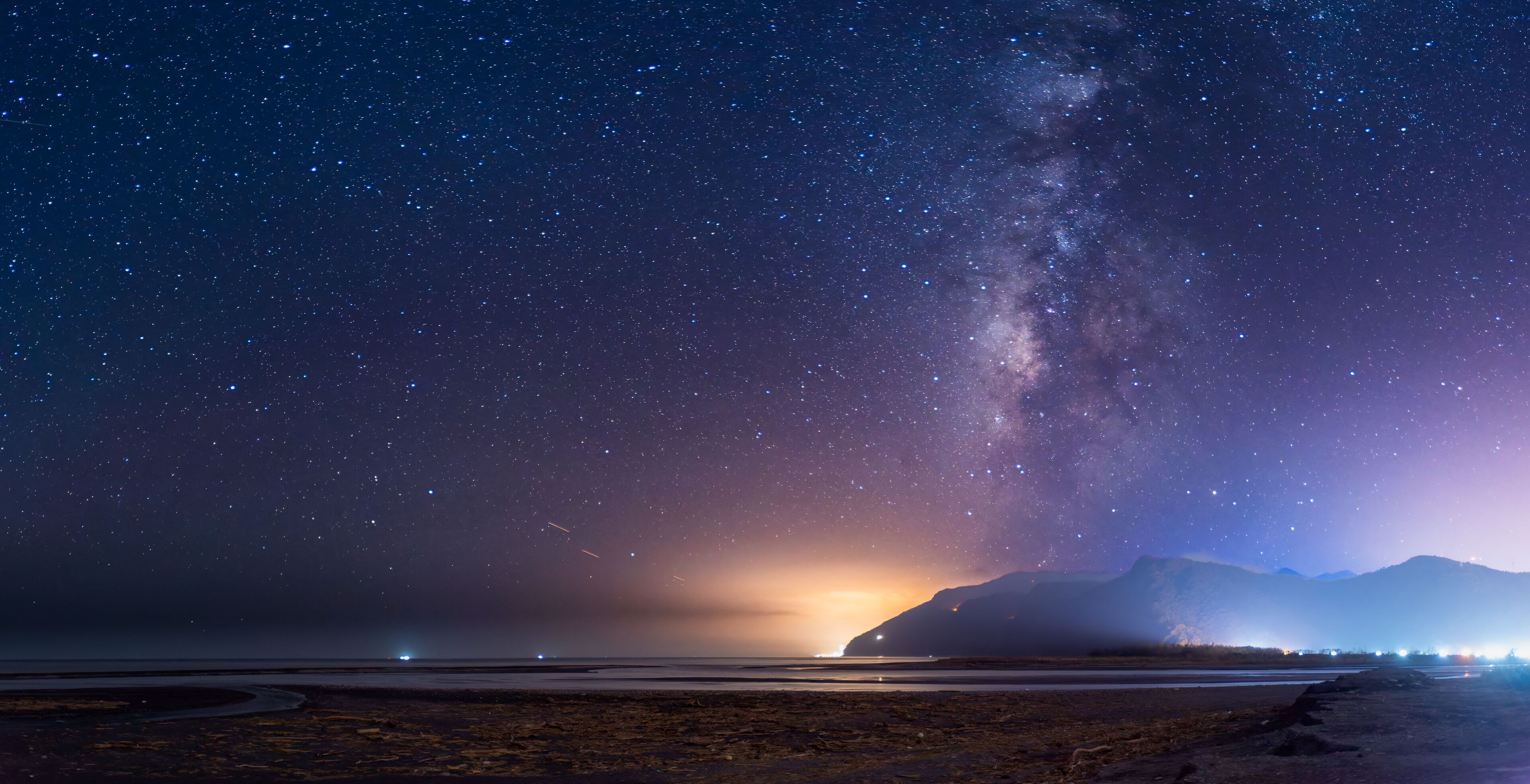 A serene night sky illuminates the coastal mountains. The Milky Way stretches across the horizon, casting a soft glow over the tranquil seascape. Suao, Yilan County, Taiwan.