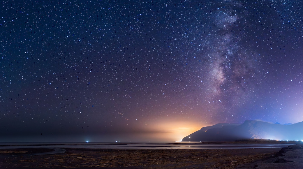 A serene night sky illuminates the coastal mountains. The Milky Way stretches across the horizon, casting a soft glow over the tranquil seascape. Suao, Yilan County, Taiwan.
